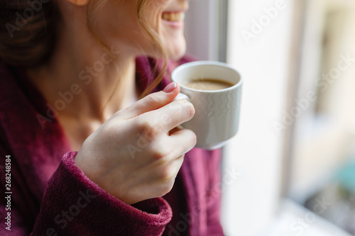 Morning coffee. Woman holding white Cup of black coffee sitting by the window. Young girl enjoys hot invigorating coffee in the morning