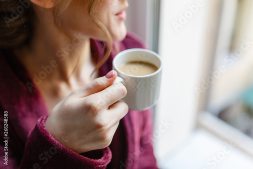 Morning coffee. Woman holding white Cup of black coffee sitting by the window. Young girl enjoys hot invigorating coffee in the morning