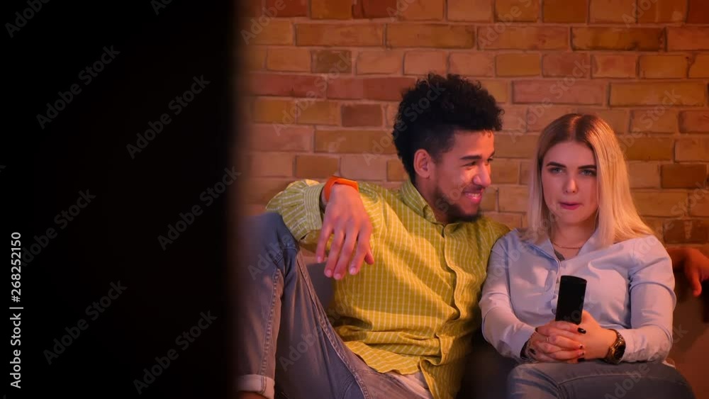 Dolly shot of young african guy with his blonde caucasian girlfriend watching TV in cozy home atmosphere. Dolly shot of young african guy with his blonde caucasian girlfriend watching TV in cozy home atmosphere.