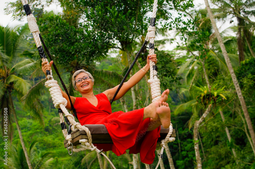 natural lifestyle portrait of attractive happy middle aged 40s - 50s Asian woman with grey hair and stylish red dress riding rainforest swing carefree swinging