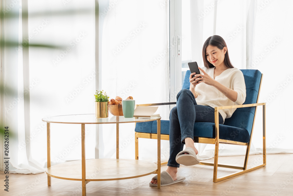 Young beautiful Asian woman relaxing in living room at home, reading books, drinking coffee and using phone in the morning with happy feeling Young beautiful Asian woman relaxing in living room at home, reading books, drinking coffee and using phone in the morning with happy feeling