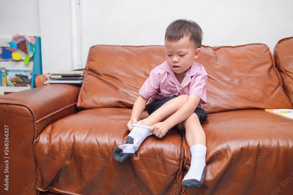 Cute little Asian 2 - 3 years old toddler boy child sitting on sofa in living room concentrate on putting on his own socks, Encourage Self-Help Skills in Children, Develop Confidence concept