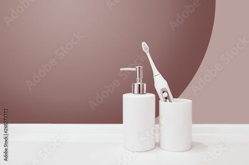 White ceramic bathroom accessories dispenser for soap and a glass with a tooth electric brush isolated on purple pastel background. copy space blank