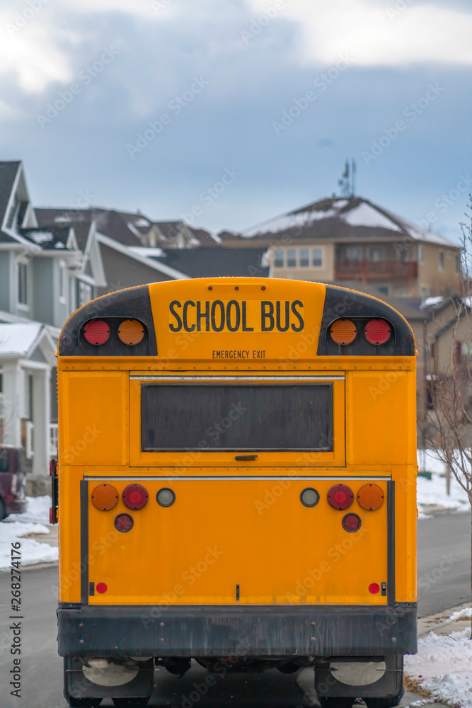 Rear of a yellow school bus against snowy homes and cloudy sky in winter