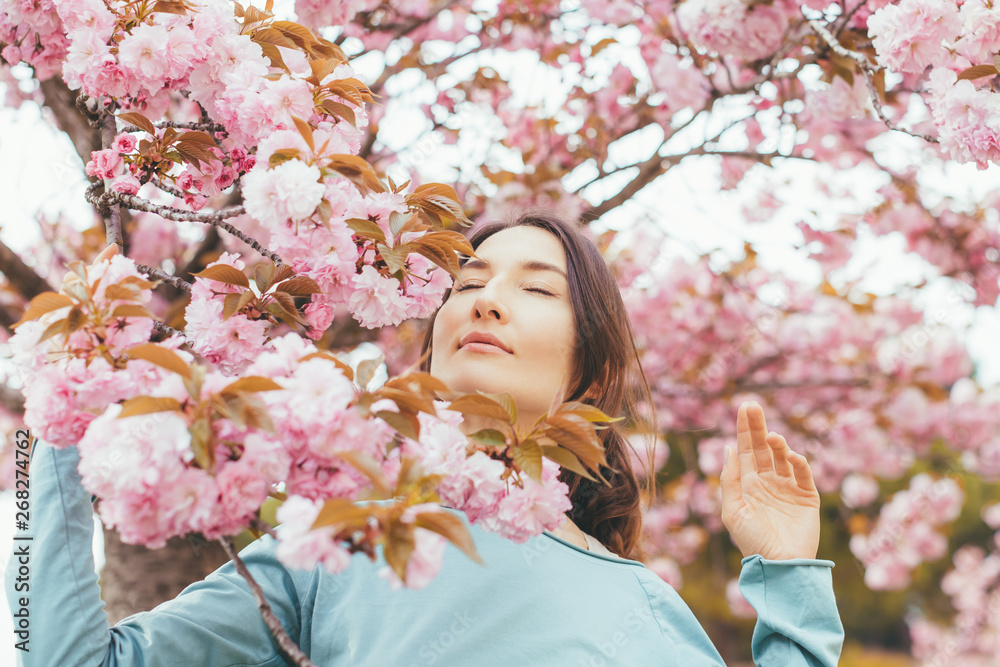Portrait of beautiful brunette woman with blooming Japanese cherry tree (Prunus serrulata) - girl inhales the scent of flowers with closed eyes - spring, nature and beauty concept Portrait of beautiful brunette woman with blooming Japanese cherry tree (Prunus serrulata) - girl inhales the scent of flowers with closed eyes - spring, nature and beauty concept