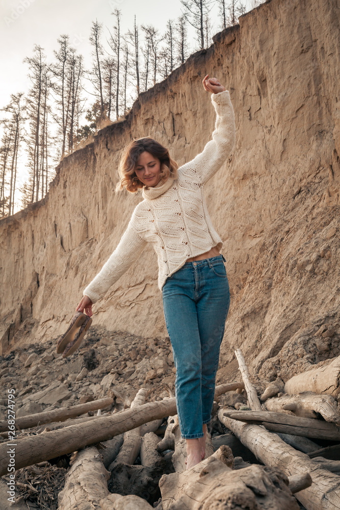 Fototapeta premium portrait of young trendy woman dressed in brown knit sweater made of natural wool and jeans laughing, smiling, posing n the beach; in the background sandy slope with trees.