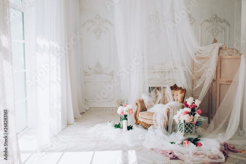 vintage armchair decorated with peonies  flowers and greens, stands in a classic room on a white wooden floor surrounded by candles near large window and curtains