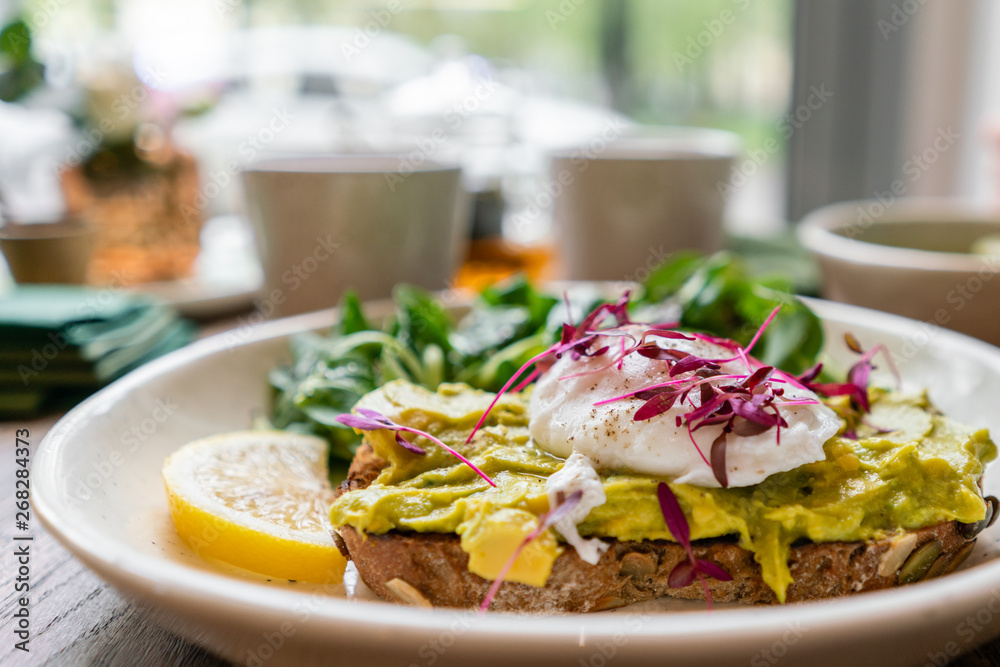 Morning in cafe, oak table. Healthy breakfast with wholemeal bread toast with avocado, poached egg with green salad. Green tea on the Background. Morning in cafe, oak table. Healthy breakfast with wholemeal bread toast with avocado, poached egg with green salad. Green tea on the Background.