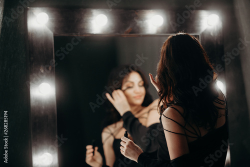 beautiful elegant brunette in a black silk dressing gown is standing in front of a tall make-up mirror and dressing herself up, paints eyelashes with mascara in a gray luxurious bathroom