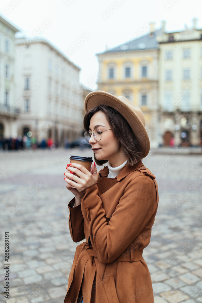 Fototapeta premium Young stylish woman in hat and coat drinking coffee to go in a city street