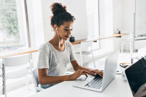 Romantic african girl with trendy hairstyle sitting at her workplace and analysing data. Indoor portrait of black female student working with laptop before exam.