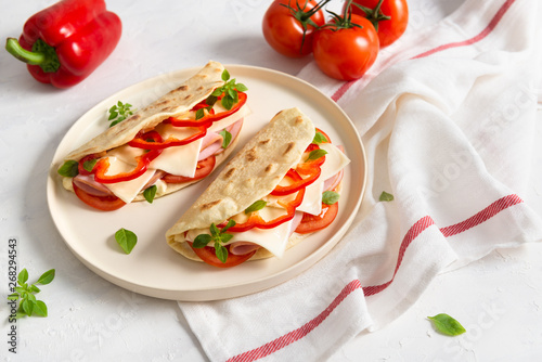 Italian piadina romagnola flatbread with red pepper, tomatoes, prosciutto ham, cheese and basil on the plate on white wooden background. Italian restaurant cuisine. Copy space