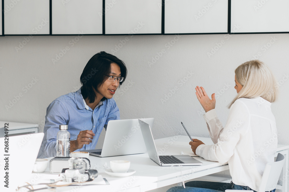 Cheerful asian student speaking with blonde girl while sitting at the table with laptop and glass of water. International freelance specialists sharing work experience with each other. Cheerful asian student speaking with blonde girl while sitting at the table with laptop and glass of water. International freelance specialists sharing work experience with each other.