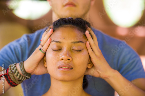 natural lifestyle portrait of young beautiful and relaxed Asian Balinese woman receiving a healing facial and head Thai massage by male therapist at traditional spa