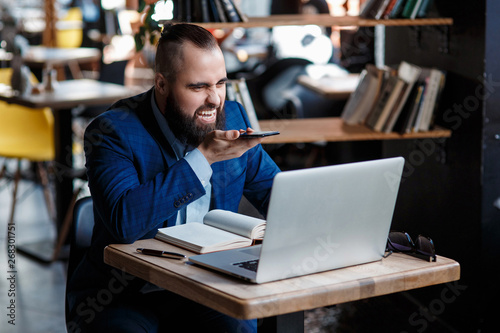 Serious bearded businessman conducts telephone conversations on a mobile phone at the computer. Unhappy man scolds on the phone. Emitations of rage in the office at the manager