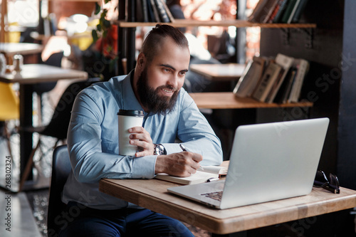 Wallpaper Mural Young serious bearded businessman working on computer at table,drinking coffee.Man analyzes information, data, develops business plan. Freelancer, entrepreneur.Online marketing, education, e-learning Torontodigital.ca