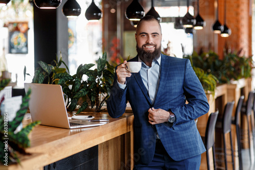 Young serious bearded businessman working on computer at table,drinking coffee.Man analyzes information, data, develops business plan. Freelancer, entrepreneur.Online marketing, education, e-learning