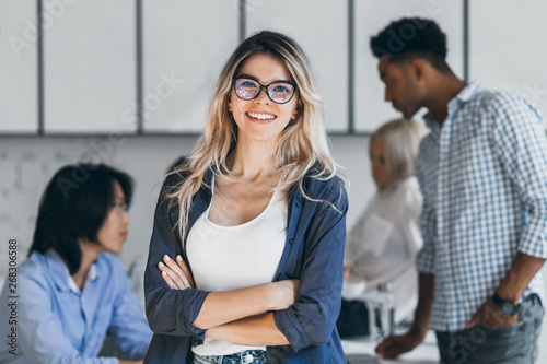 Confident blonde female manager posing with smile after conference with other employees. Asian programmer talking with african freelancer while fair-haired secretary laughing on foreground. photo