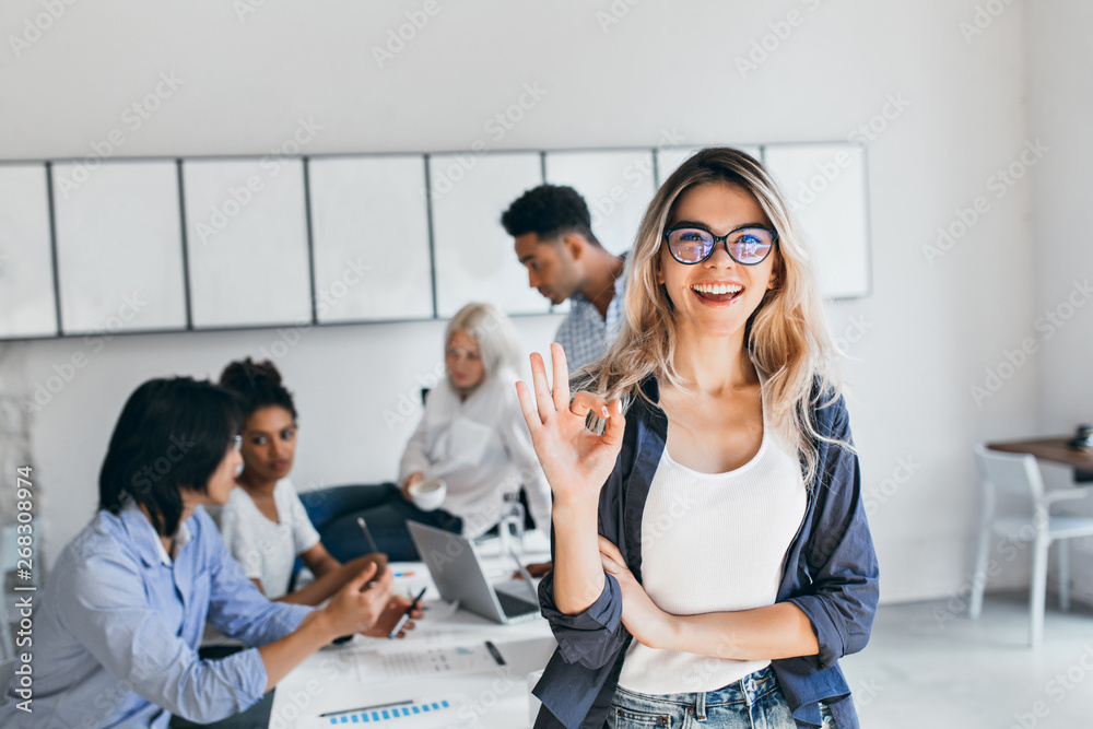 African students with serious face expression preparing for seminar while blonde girl posing with okay sign. Young female businesswoman having fun in office after brainstorm with colleagues. - obrazy, fototapety, plakaty 