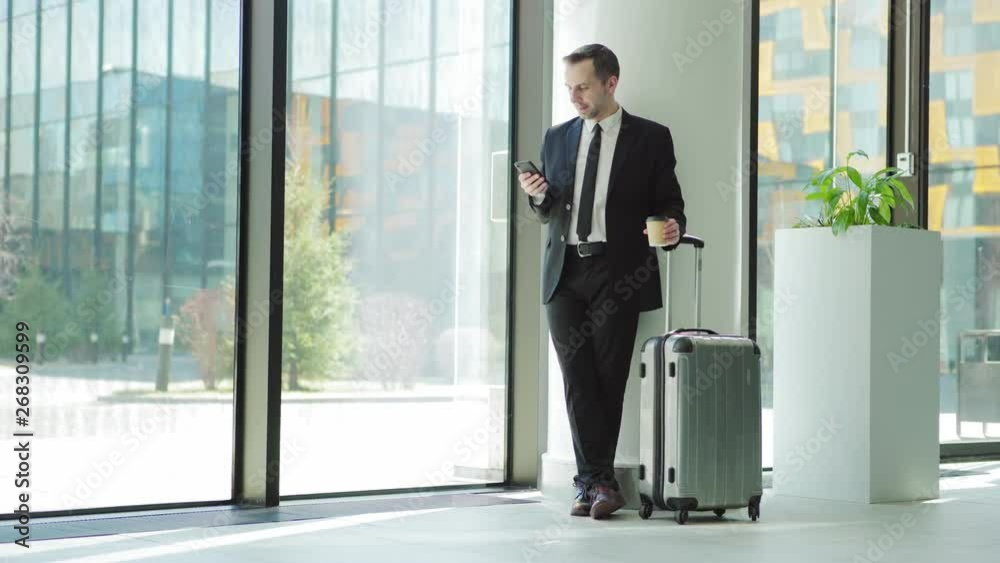Wide shot of middle aged businessman text messaging on cell phone and drinking coffee while leaning on column in airport and waiting for flight with his trolley suitcase Wide shot of middle aged businessman text messaging on cell phone and drinking coffee while leaning on column in airport and waiting for flight with his trolley suitcase