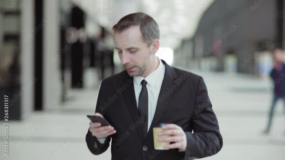 Dolly medium shot of confident executive in suit and necktie using cell phone while walking in hall of business center, takeaway coffee cup in his hand Dolly medium shot of confident executive in suit and necktie using cell phone while walking in hall of business center, takeaway coffee cup in his hand