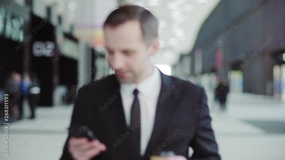 Dolly medium shot of confident middle aged manager in suit and necktie typing messages on cell phone and drinking takeaway coffee while walking along hall in business center, mall or airport Dolly medium shot of confident middle aged manager in suit and necktie typing messages on cell phone and drinking takeaway coffee while walking along hall in business center, mall or airport