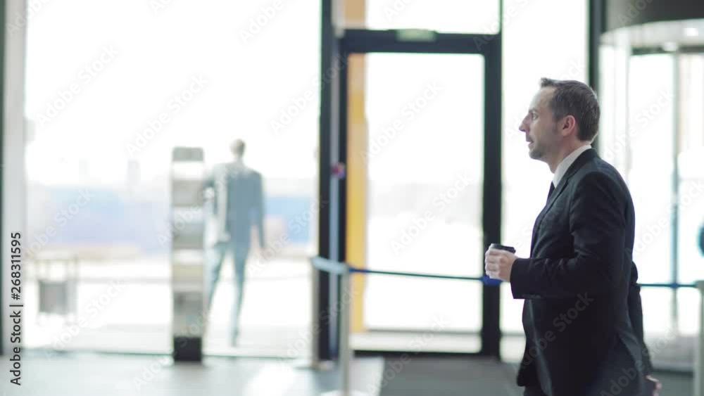 Side view tracking left shot of confident businessman in formal suit walking in entrance hall of business center, mall or airport; takeaway coffee cup and cell phone in his hands Side view tracking left shot of confident businessman in formal suit walking in entrance hall of business center, mall or airport; takeaway coffee cup and cell phone in his hands
