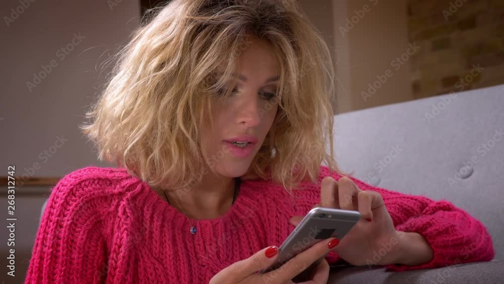 Close-up shot pretty housewife in pink sweater surfing attentively in smartphone shows surprise in home atmosphere. Close-up shot pretty housewife in pink sweater surfing attentively in smartphone shows surprise in home atmosphere.