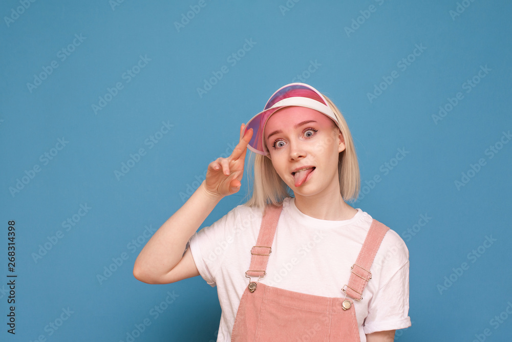 Portrait of a funny girl in a pretty casual dress, wearing a pink cap and making a funny face on a blue background. Funny teen girl poses on the camera, isolated closeup portrait. Portrait of a funny girl in a pretty casual dress, wearing a pink cap and making a funny face on a blue background. Funny teen girl poses on the camera, isolated closeup portrait.