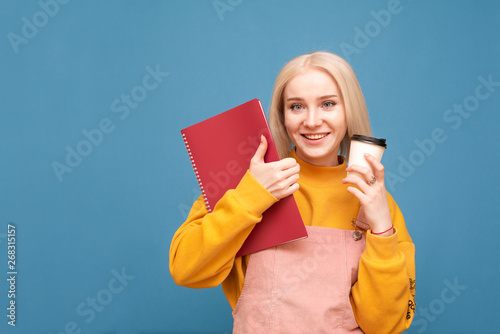 Happy blonde girl holds a cup of coffee and a notebook in her hands, looks at the camera and smiles, isolated on a blue background. Smiling student girl stands on a blue background and rejoices.