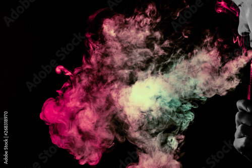 Side part of guys gray face and vape in arm with a colored backlight of monochrome smoking vape and exhaling pink  green and white in different direction on a black background. Puffs harmful to health