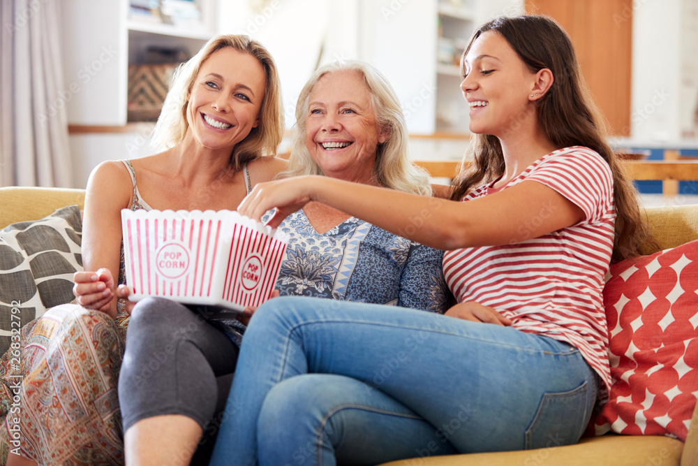 Mother With Adult Daughter And Teenage Granddaughter Eating Popcorn Watching Movie On Sofa At Home