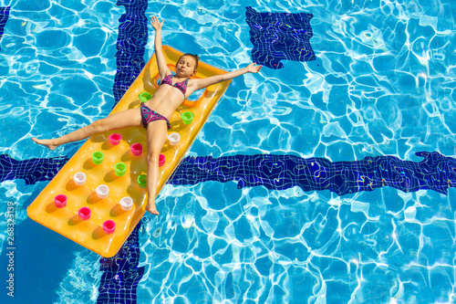 A little girl sunbathing on a floating orange air mattress in the pool.