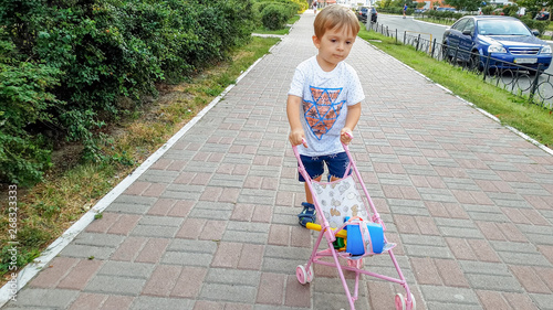 3 years old toddler boy walking with toy pram on the street. Boy playing with toys for girls.