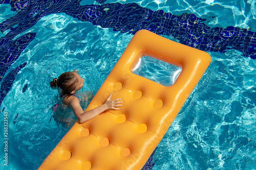 A little girl is swimming with an orange air mattress in the pool on a Sunny day.