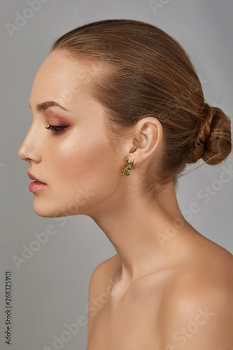 Cropped side view shot of woman with chestnut hair, tied back in a bun, with naked chest and fresh makeup, looking down. The girl with tanned skin is wearing bronze earring in the shape of Pegasus.