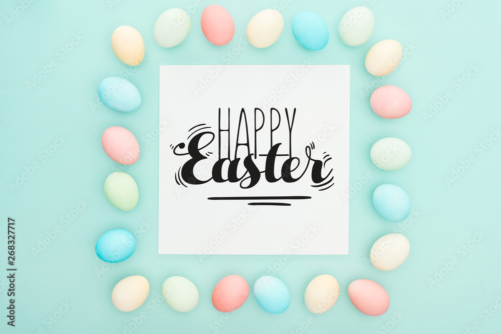 top view of square frame made of painted chicken eggs on blue background with happy Easter black lettering on white greeting card