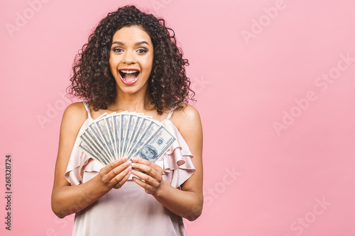 Rich girl! Money winner! Surprised beautiful african american woman in dress holding money and looking at the camera isolated against pink background.