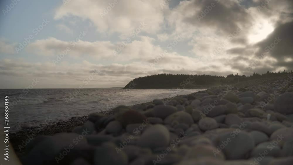 Ground shot of some waves hitting beach with some stones on the ground, trees and a road on the background near sunset in Halifax, Nova Scotia, Canada. Ground shot of some waves hitting beach with some stones on the ground, trees and a road on the background near sunset in Halifax, Nova Scotia, Canada.