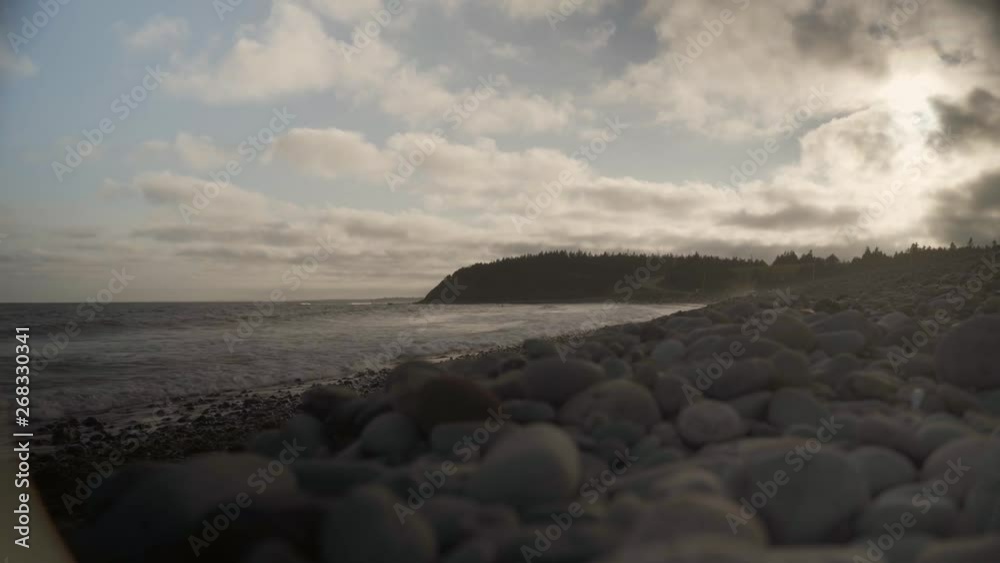Ground shot of some waves hitting beach with some stones on the ground, trees and a road on the background near sunset in Halifax, Nova Scotia, Canada.