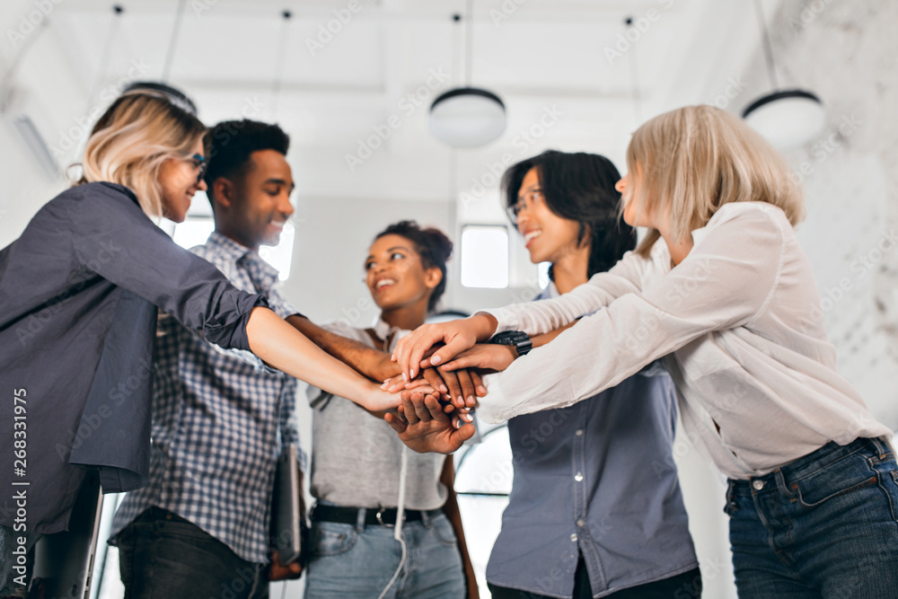 Cheerful international students with happy face expression going to work together on science project. Indoor photo of blonde woman in trendy blouse holding hands with coworkers. Cheerful international students with happy face expression going to work together on science project. Indoor photo of blonde woman in trendy blouse holding hands with coworkers.