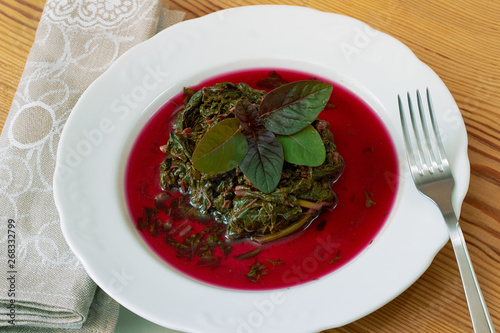 Prepaired braised amaranth - red spinach, in it's own sauce and a fresh leaf on top on white plate on wooden table  with linen napkin and fork nearby, copy space, close up, top view photo