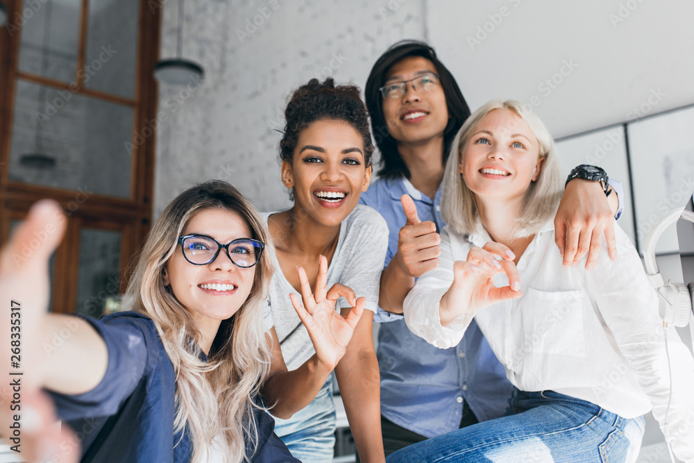 Fototapeta premium Young international office workers posing together and laughing during break. Indoor portrait of excited european blonde girl in glasses having fun with colleagues.