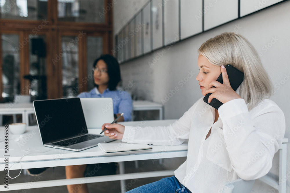 Lovely blonde girl in white shirt calling boss to make report while sitting in office. Indoor portrait of female manager with serious face expression with her asian male co-worker. Lovely blonde girl in white shirt calling boss to make report while sitting in office. Indoor portrait of female manager with serious face expression with her asian male co-worker.