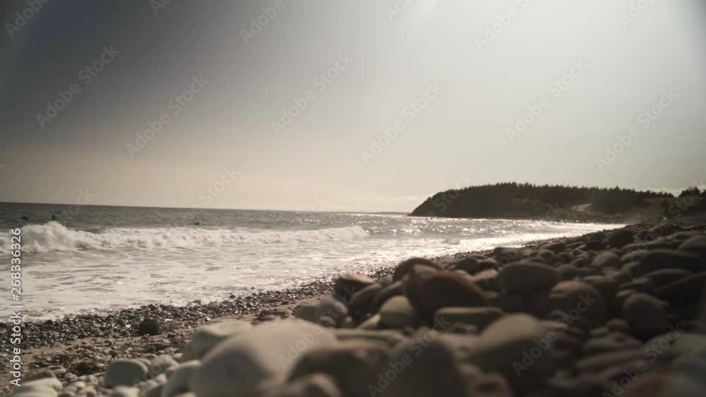 Ground shot of some waves hitting beach with some stones on the ground, trees and a road on the background near sunset in Halifax, Nova Scotia, Canada. Ground shot of some waves hitting beach with some stones on the ground, trees and a road on the background near sunset in Halifax, Nova Scotia, Canada.