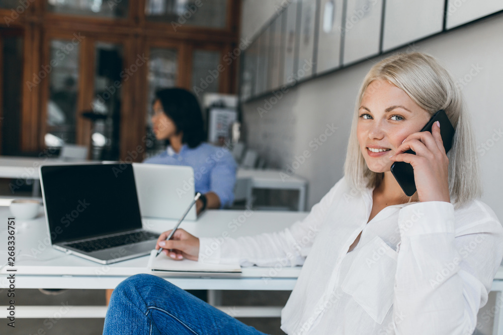 Cute blonde secretary in white blouse talking on phone and writing data in notebook. Indoor portrait of long-haired asian it-specialist with graceful lady at reception desk. Cute blonde secretary in white blouse talking on phone and writing data in notebook. Indoor portrait of long-haired asian it-specialist with graceful lady at reception desk.