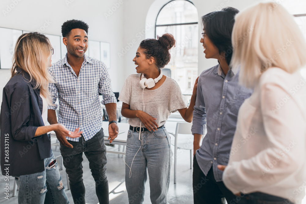 Surprised african student in blue shirt telling friends about exams. Indoor portrait of black curly girl in jeans spending time with freelance colleagues in office. Surprised african student in blue shirt telling friends about exams. Indoor portrait of black curly girl in jeans spending time with freelance colleagues in office.