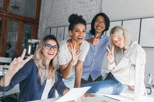 Two blonde female office workers posing with peace sign while fooling around with foreign colleagues. Short-haired african it-specialist having fun at workplace with asian friend. photo