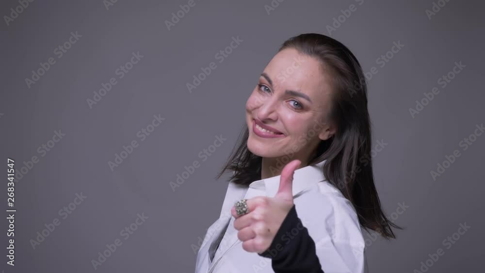 Closeup portrait of adult attractive caucasian female turning around with her hair fluttering and happily showing thumb up looking at camera with background isolated on gray Closeup portrait of adult attractive caucasian female turning around with her hair fluttering and happily showing thumb up looking at camera with background isolated on gray