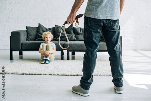 partial view of abusive father holding belt and sad son sitting on carpet